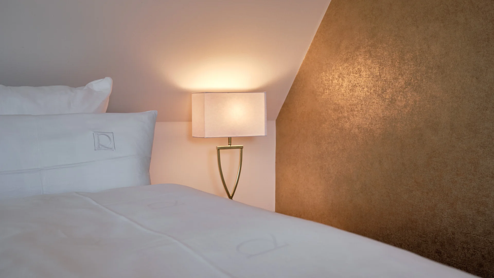 Reppert, Ihr Hotel in Hinterzarten im Schwarzwald Weißes Bett mit Kissen neben einer leuchtenden Nachttischlampe und goldenem Wandpaneel