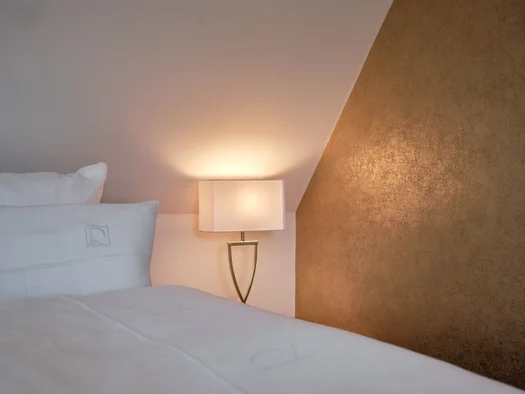 Reppert, Ihr Hotel in Hinterzarten im Schwarzwald Weißes Bett mit Kissen neben einer leuchtenden Nachttischlampe und goldenem Wandpaneel
