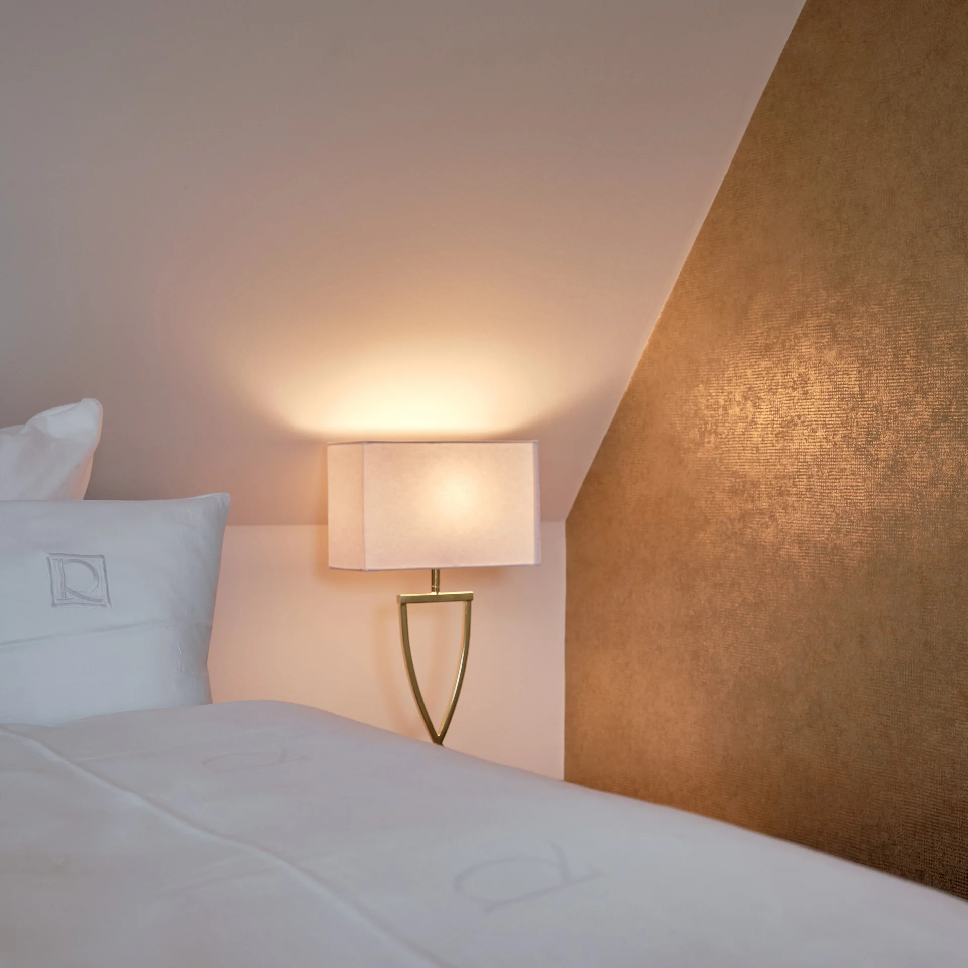 Reppert, Ihr Hotel in Hinterzarten im Schwarzwald Weißes Bett mit Kissen neben einer leuchtenden Nachttischlampe und goldenem Wandpaneel